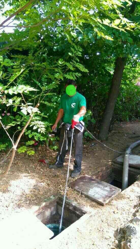 Un ouvrier en chemise verte actionne une pompe près d'une fosse à ciel ouvert, à l'extérieur, sous des arbres.