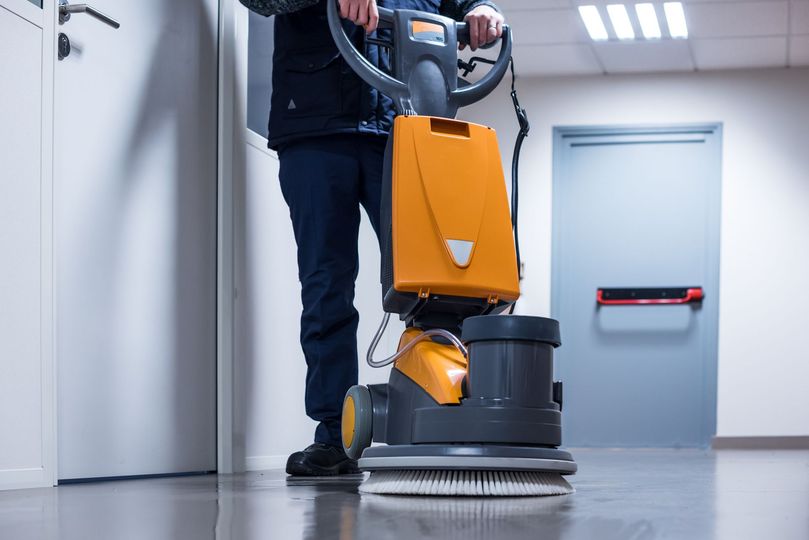 Une personne utilise une machine de nettoyage de sol orange et grise dans un couloir.