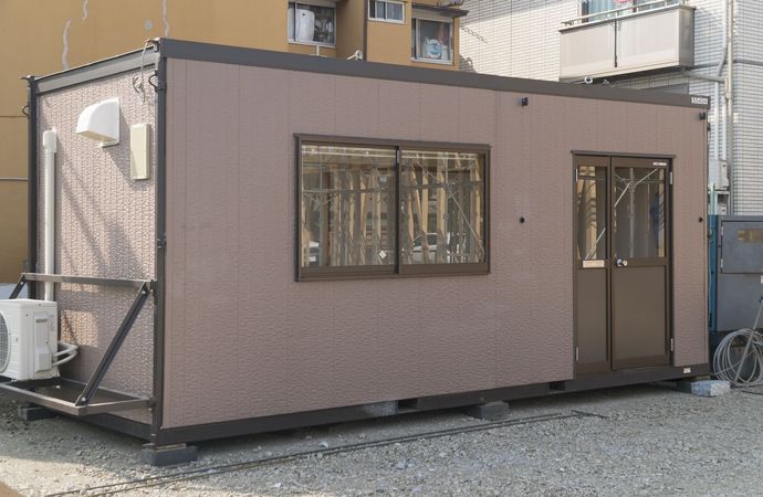 Bâtiment préfabriqué rectangulaire brun avec fenêtre, porte et unité de climatisation.