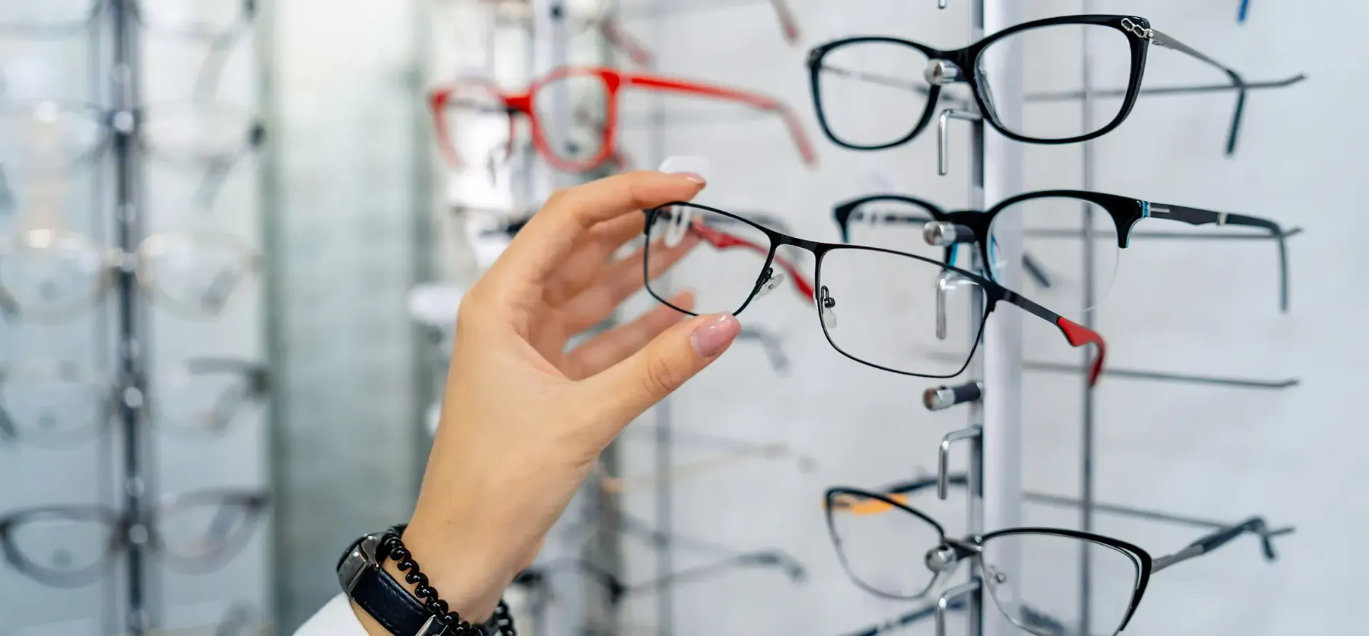 In einem Optikergeschäft steht eine Person mit verschränkten Armen; im verschwommenen Hintergrund arbeitet ein Kollege an einem Computer.