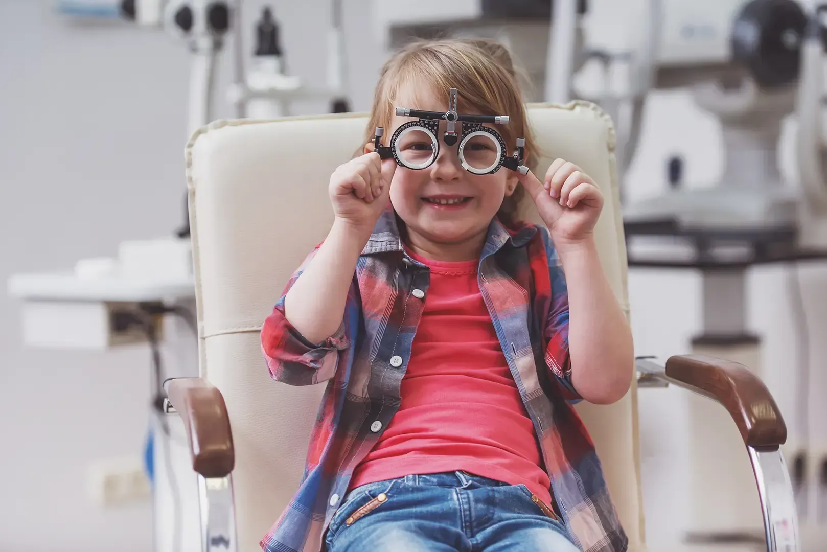 Ein lächelndes Kind, das eine verstellbare Probebrille für eine Augenuntersuchung trägt, sitzt auf einem Untersuchungsstuhl in der Praxis eines Optikers.