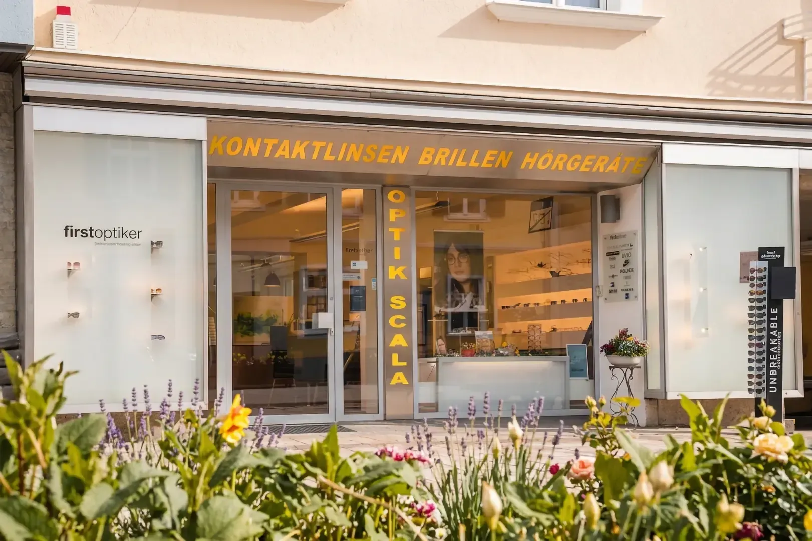 Vier Brillenträger stehen nebeneinander in einem Optikergeschäft; im Hintergrund sind Regale mit Brillengestellen ausgestellt.