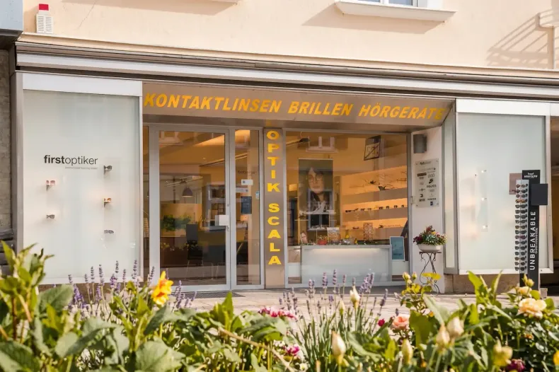 Vier Brillenträger stehen nebeneinander in einem Optikergeschäft; im Hintergrund sind Regale mit Brillengestellen ausgestellt.