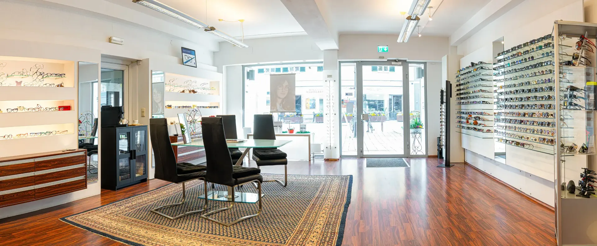 Heller Brillen-Showroom mit Ausstellungsständern, Stühlen, gemustertem Teppich und gläserner Fronttür