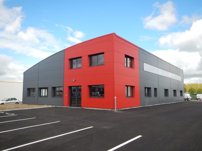 Bâtiment industriel moderne avec façade rouge et grise, fenêtres noires et parking pavé.