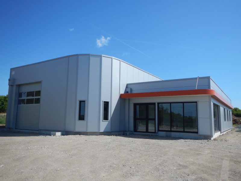Bâtiment commercial gris avec des boiseries orange et une grande porte de garage. Ciel bleu dégagé.