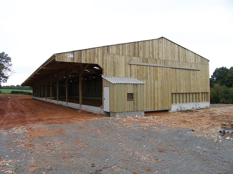 Grande grange en bois avec un appentis et une petite dépendance, située sur un terrain de gravier et de terre.