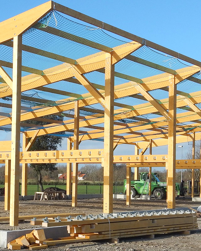Charpente en bois d'un bâtiment en construction, filet bleu, ciel lumineux, engins de chantier visibles.