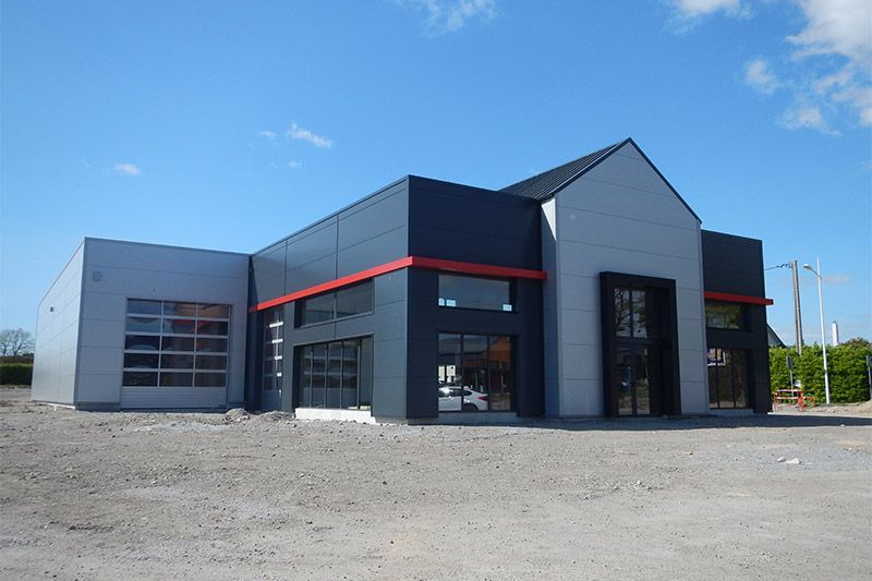 Bâtiment commercial moderne avec façade grise et noire, finitions rouges et portes/fenêtres vitrées sous un ciel bleu.