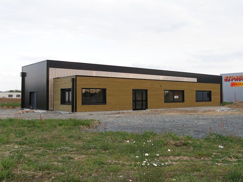 Un bâtiment moderne de plain-pied avec une façade noire et beige, des fenêtres rectangulaires et un terrain gravillonné.