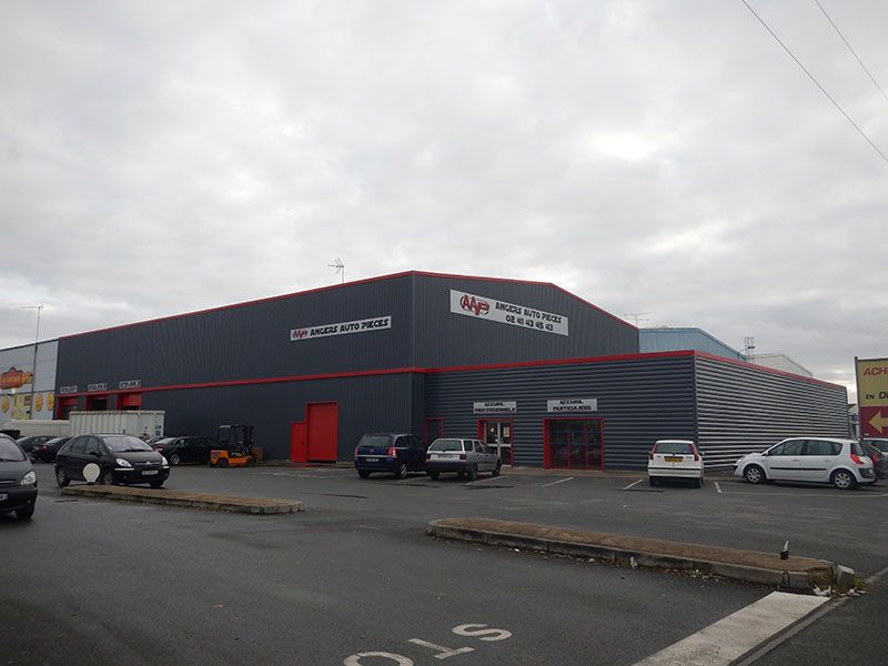 Extérieur d'un grand bâtiment gris et rouge, avec des voitures garées par temps nuageux.