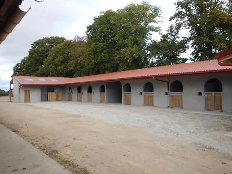 Une rangée d'écuries avec des portes en bois et des ouvertures cintrées, sous un toit de tuiles rouges, et une cour de gravier.