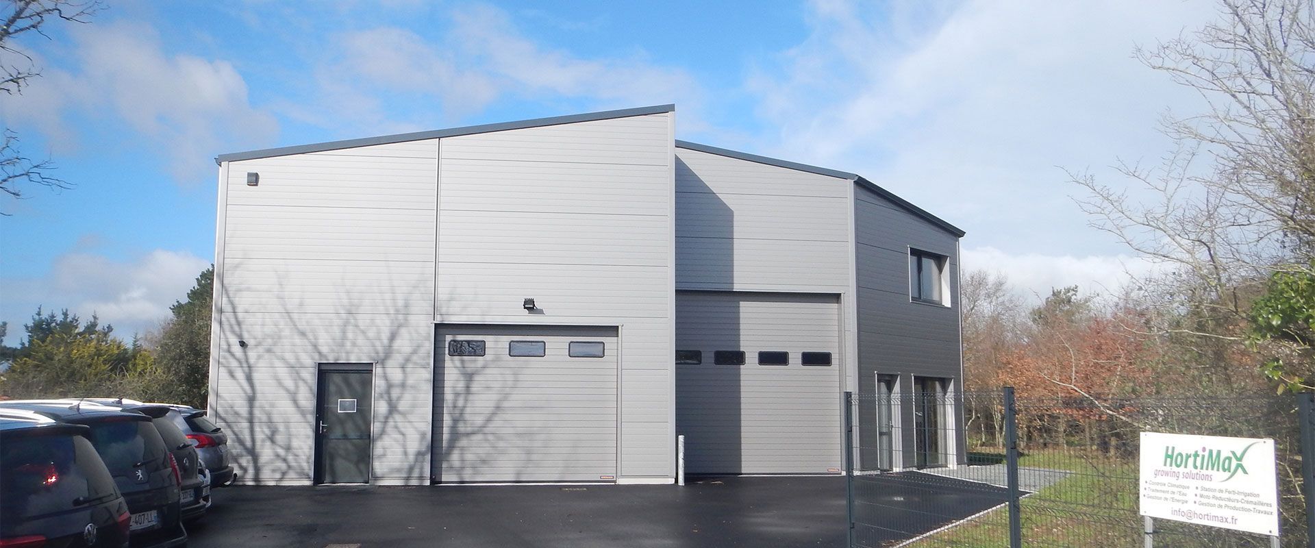 Bâtiment commercial gris moderne avec deux portes de garage et une enseigne en façade. Ciel bleu et quelques voitures.