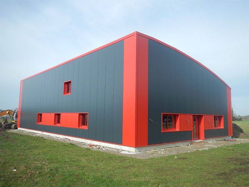 Un bâtiment industriel moderne aux murs noirs et aux encadrements de fenêtres rouges, se dresse sur un champ herbeux.