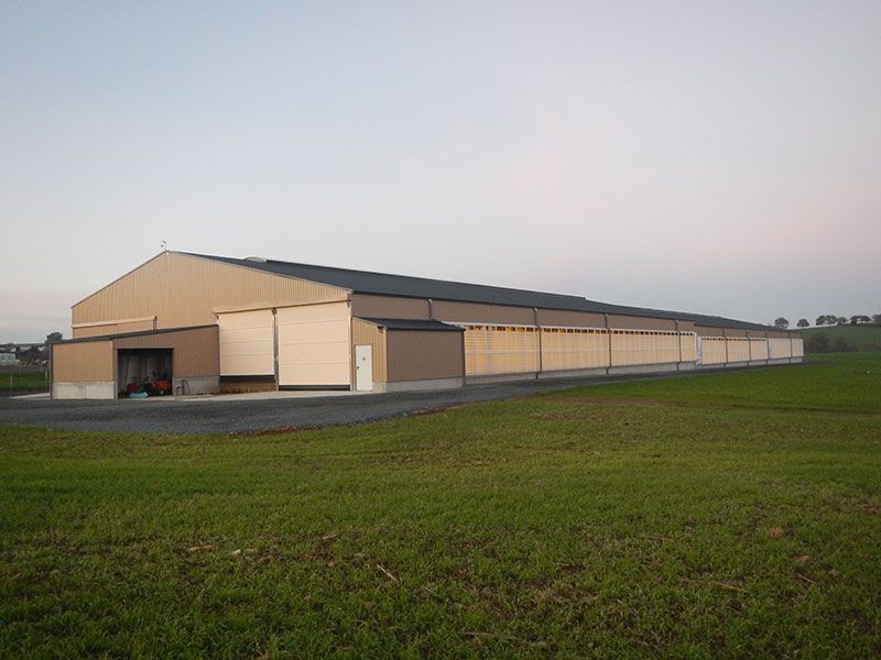 Long bâtiment métallique beige au toit sombre, situé au milieu d'un champ herbeux.