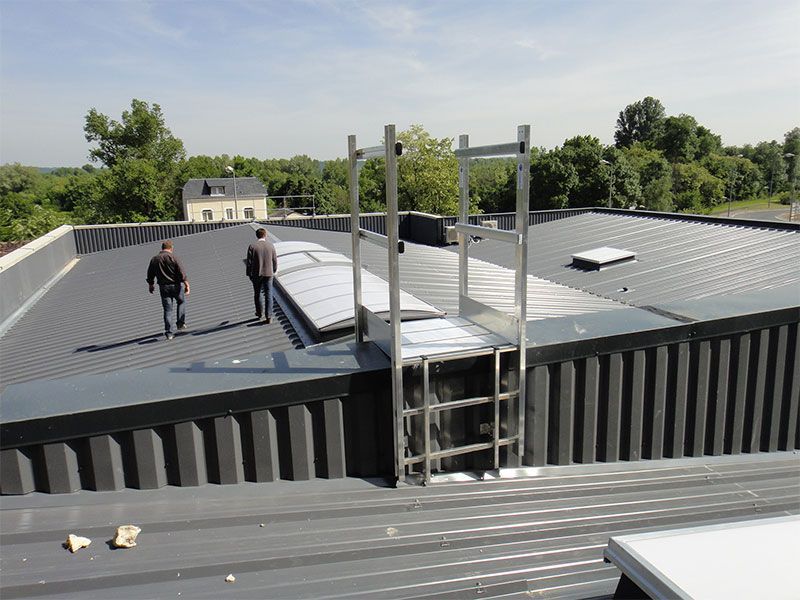 Deux personnes marchent sur un toit près d'une échelle métallique. Le toit est gris et comporte plusieurs puits de lumière.