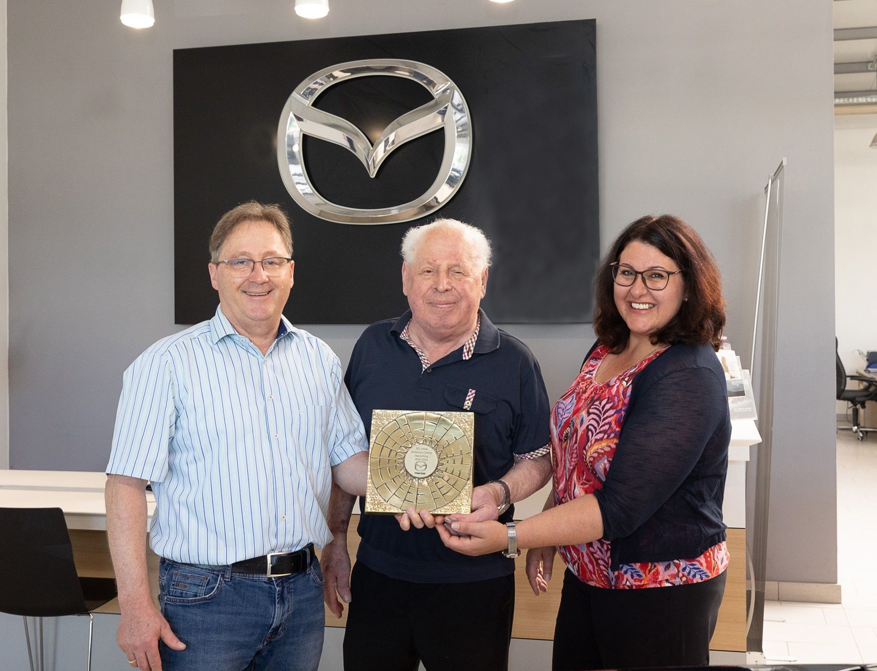 3 Personen mit einer Auszeichnung vor einem Mazda Schild an der Wand