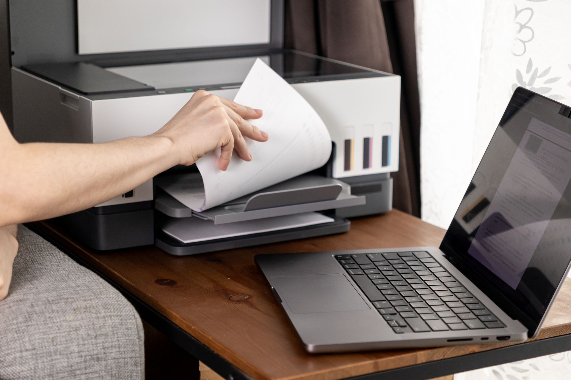 Une main place une feuille de papier dans une imprimante posée à côté d'un ordinateur portable sur un bureau.