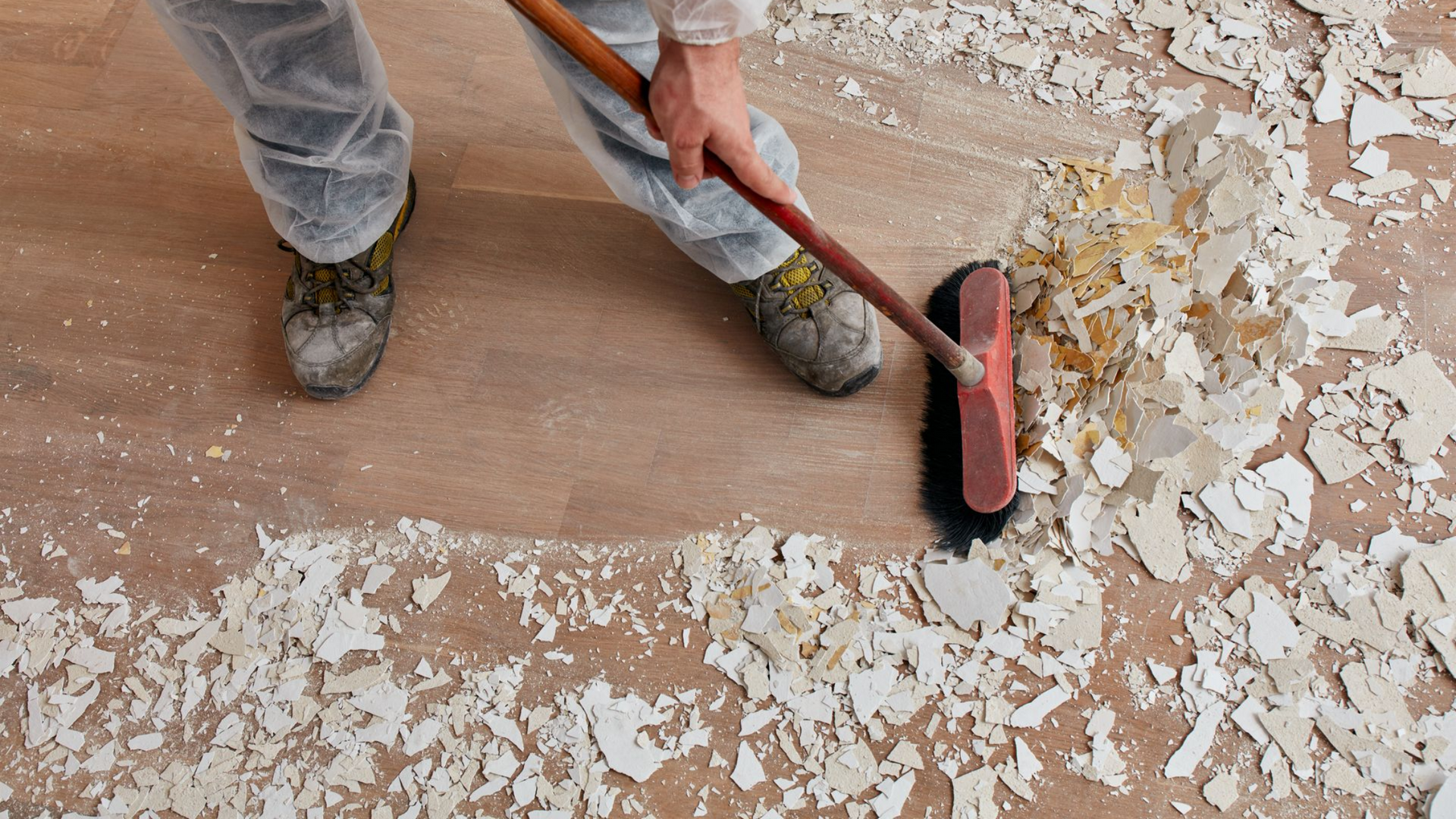 Une personne balaie les débris de construction d'un plancher en bois à l'aide d'un balai-brosse.