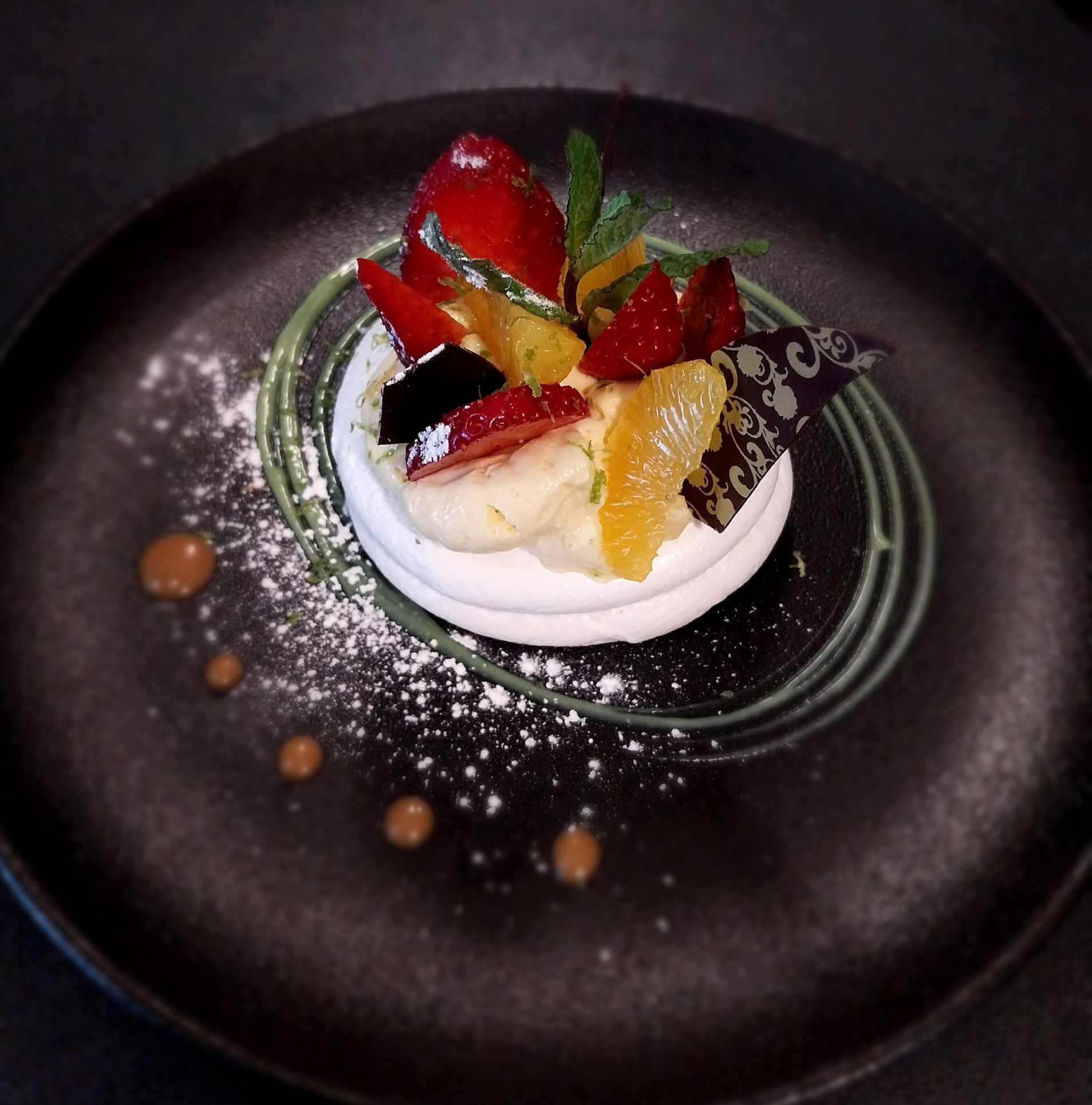 Assiette à dessert composée de meringue, de fruits rouges frais, d'agrumes et de menthe, présentée sur un plat foncé avec des points de sauce.