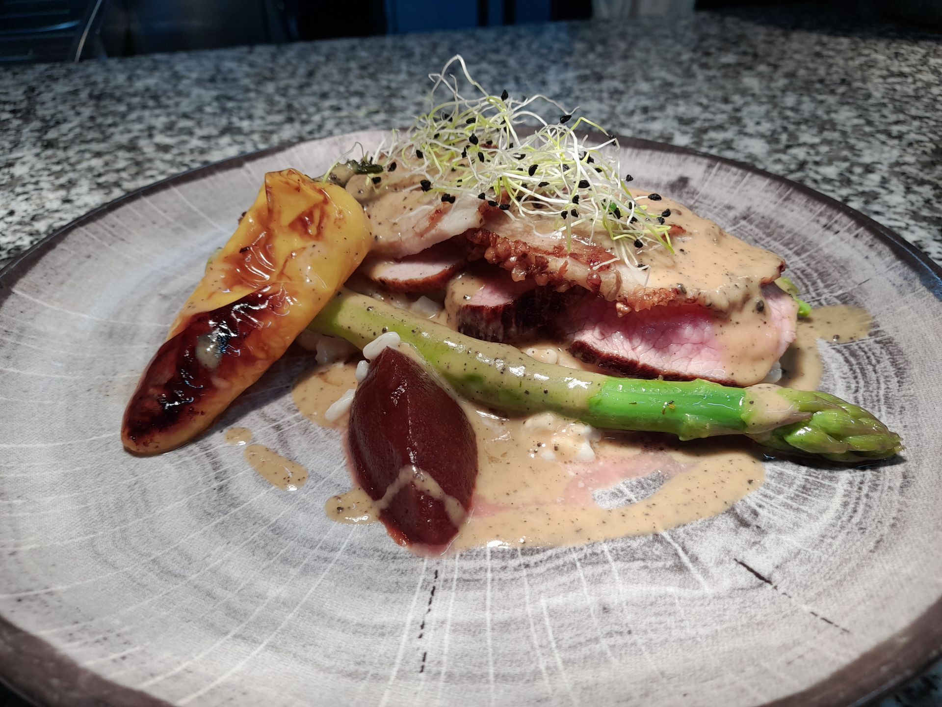 Plat gastronomique présenté dans une assiette grise, composé de viande tranchée, d'asperges, de pommes de terre rôties, de légumes verts et d'une sauce crémeuse.