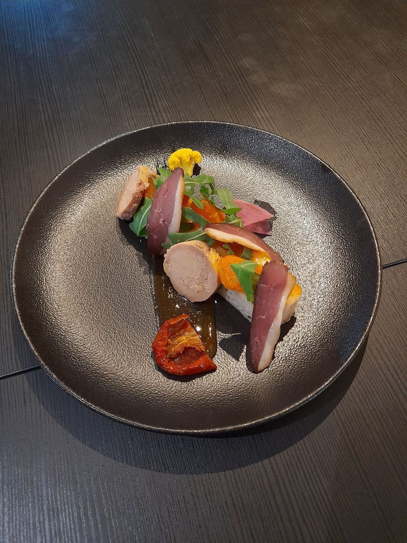 Plat gastronomique composé de viande tranchée, de légumes et de sauce, présenté sur une assiette ronde foncée.