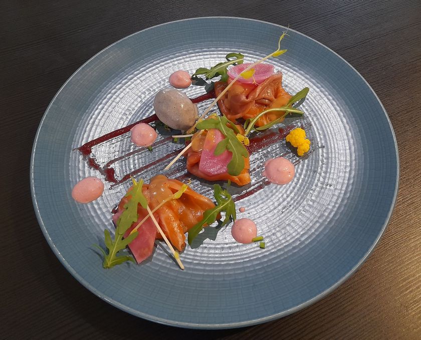Plat gastronomique composé de saumon, de légumes verts, de sauces et de fleurs comestibles, présenté sur une assiette bleue.