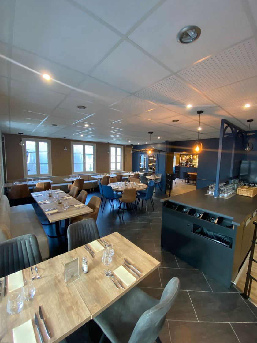Salle à manger moderne d'un restaurant avec tables et chaises en bois, grandes fenêtres et suspensions lumineuses.
