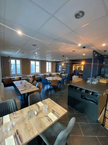 Salle à manger de restaurant vide avec de longues tables dressées, des chaises beiges et de grandes fenêtres laissant entrer la lumière du jour.