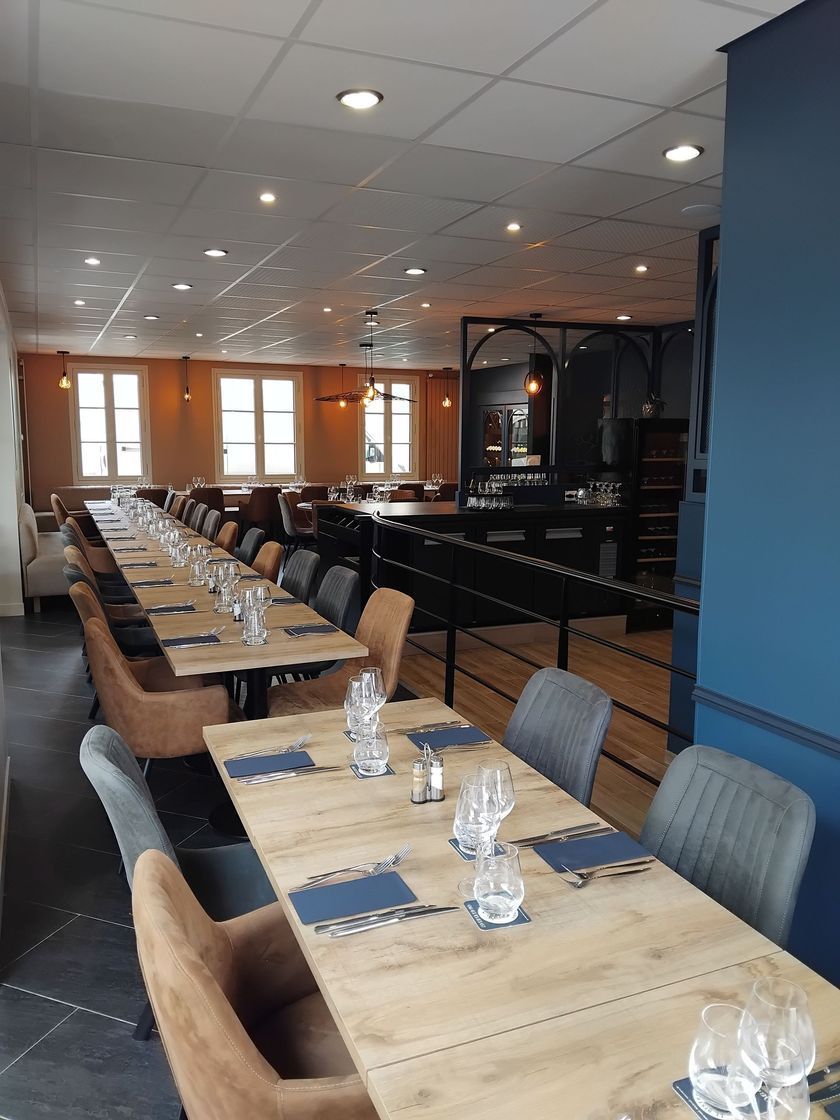 Longue salle à manger de restaurant avec tables et chaises dressées, murs bleus et luminaires suspendus.