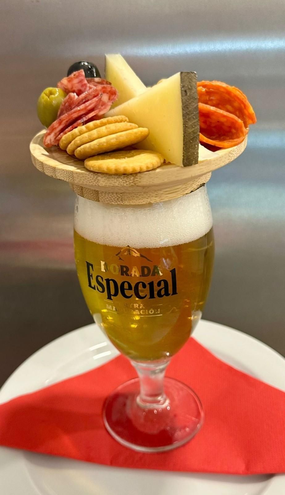 Vaso de cerveza cubierto con un plato de madera con queso, galletas, salami, aceitunas y salchichas.