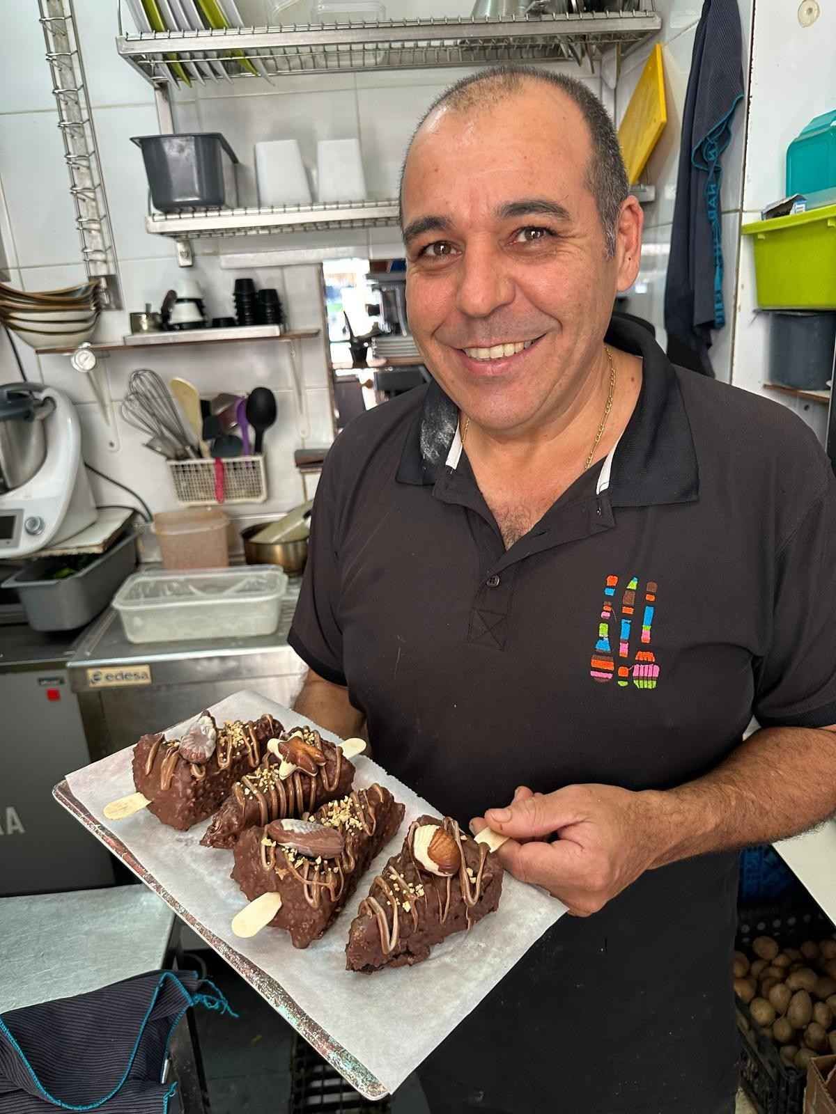 Hombre sosteniendo una bandeja de delicias cubiertas de chocolate, sonriendo en una cocina.