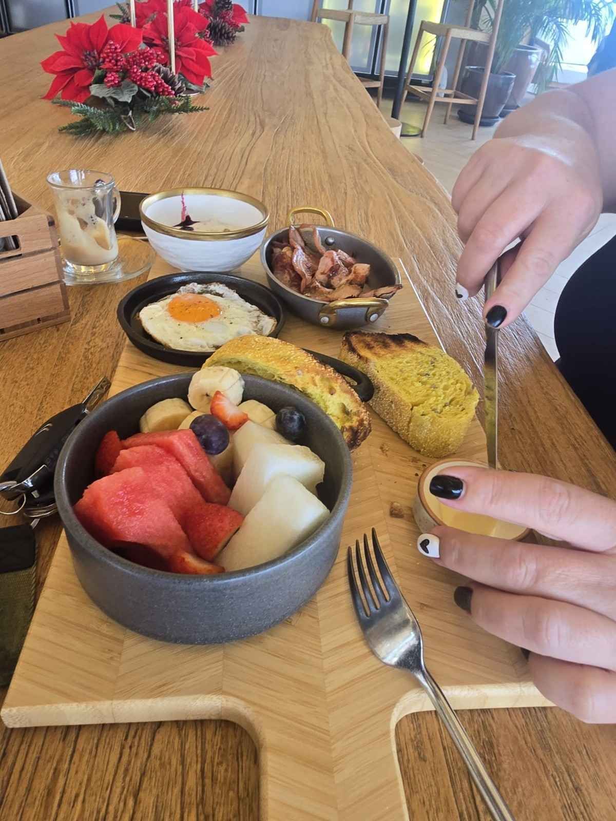 Persona cortando pan en un brunch con un plato de fruta, huevo frito, tocino y tostadas. Mesa de madera.