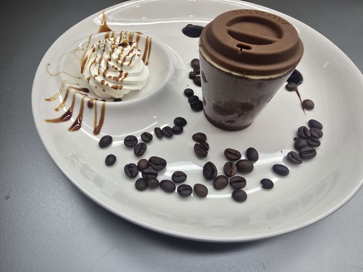 Postre de chocolate en forma de taza de café con crema batida, llovizna de chocolate y granos de café sobre un plato blanco.