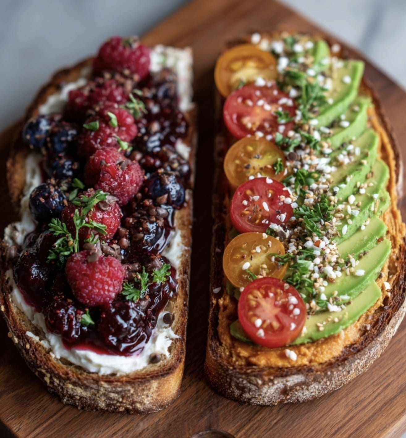 Dos rebanadas de pan tostado: una con frutos rojos y crema, la otra con aguacate y tomates, sobre una tabla de madera.