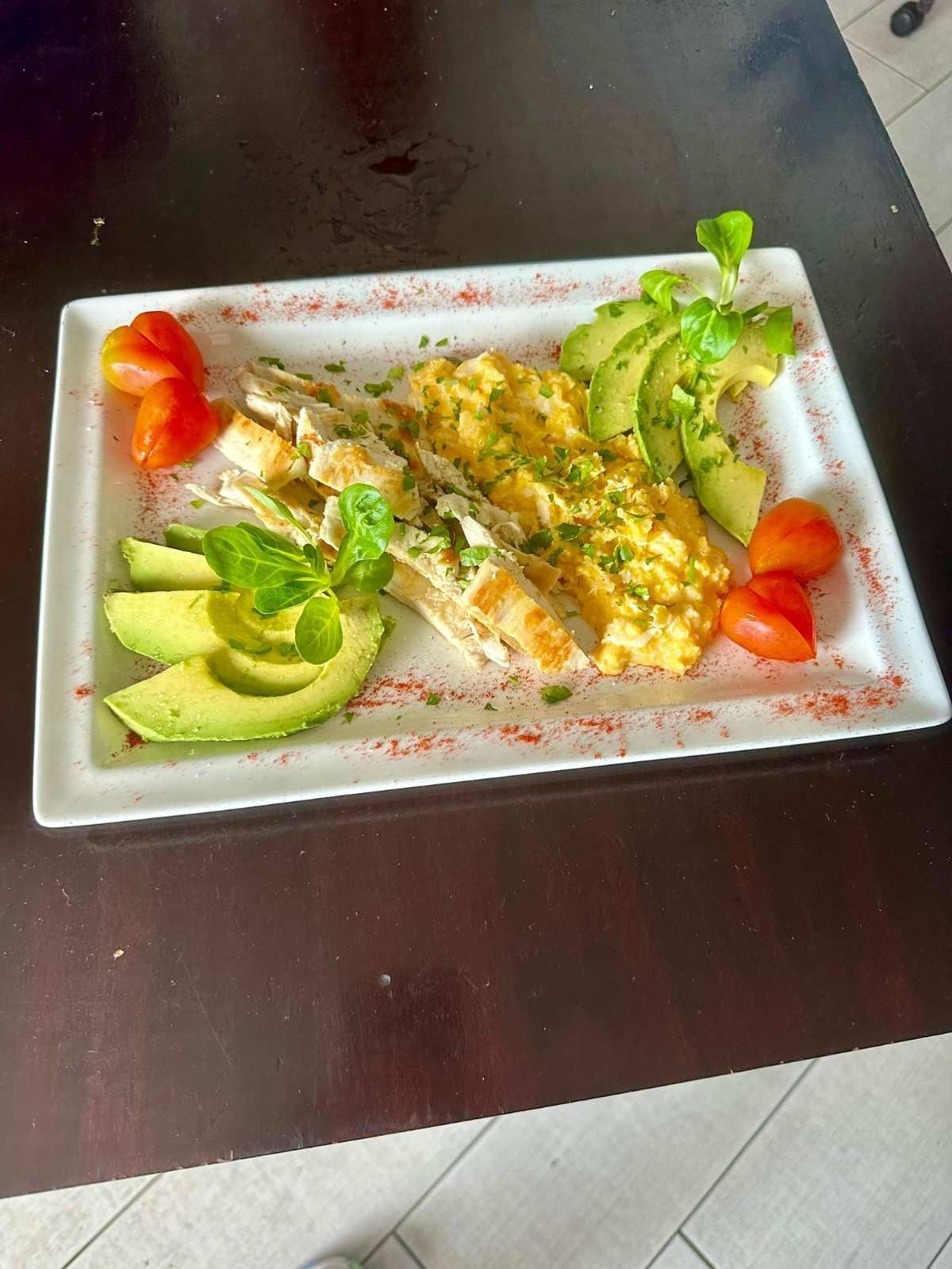 Plato de comida con rodajas de aguacate, huevos revueltos, pan y tomates, adornado con hierbas.