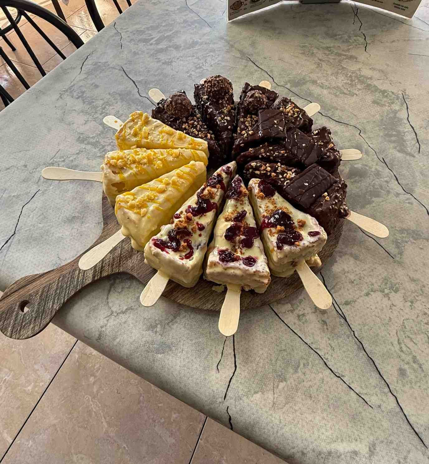 Tabla de madera con rebanadas de pastel y paletas de brownie sobre una mesa.