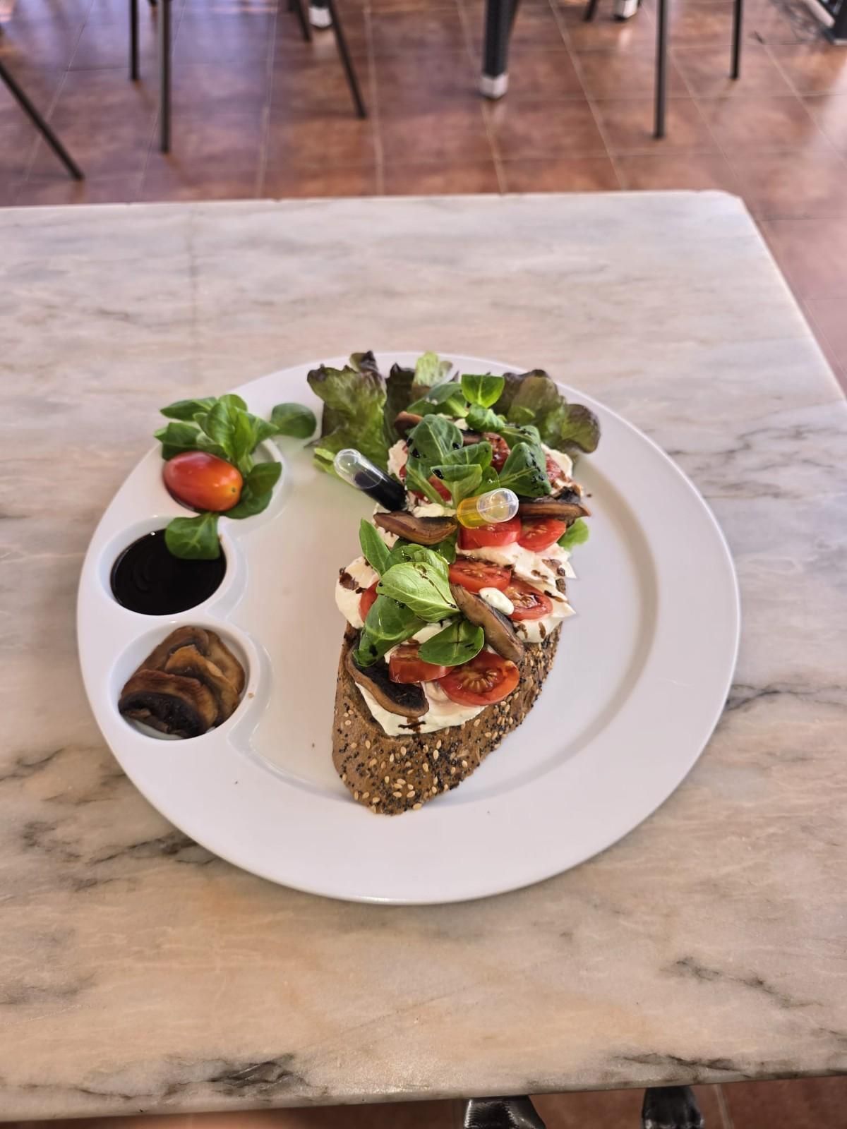 Sándwich abierto con tomates, champiñones y verduras en un plato blanco con guarniciones.