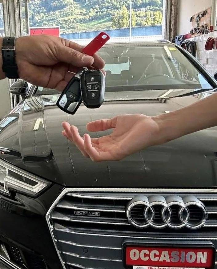 Une personne tend des clés de voiture par-dessus le capot d'une Audi noire immatriculée « OCCASION » dans un garage.