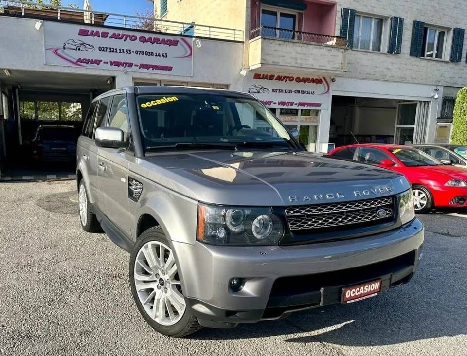 Un Land Rover Range Rover Sport gris est garé devant un garage, une voiture rouge étant visible en arrière-plan.