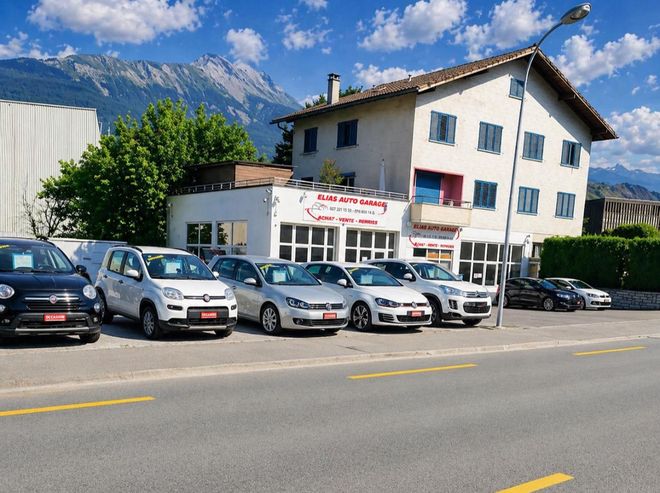 Bâtiment blanc en bord de route, avec des voitures garées devant, sur fond de montagnes et de ciel bleu nuageux.