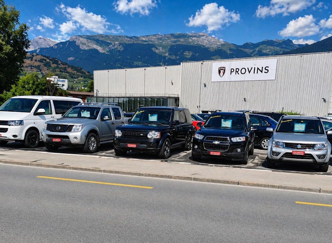 Des SUV et des pick-ups stationnés devant un bâtiment de Provins, avec des montagnes en arrière-plan sous un ciel bleu.