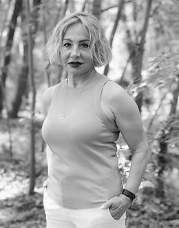 Mujer con camiseta clara y pantalones blancos en un bosque. Mira a la cámara.