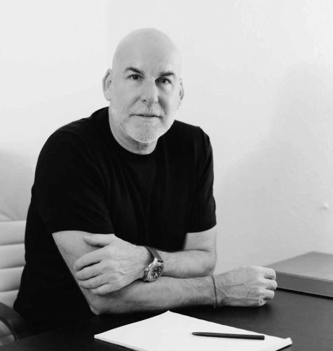 Un hombre calvo con una camisa negra está sentado en un escritorio, mirando a la cámara.