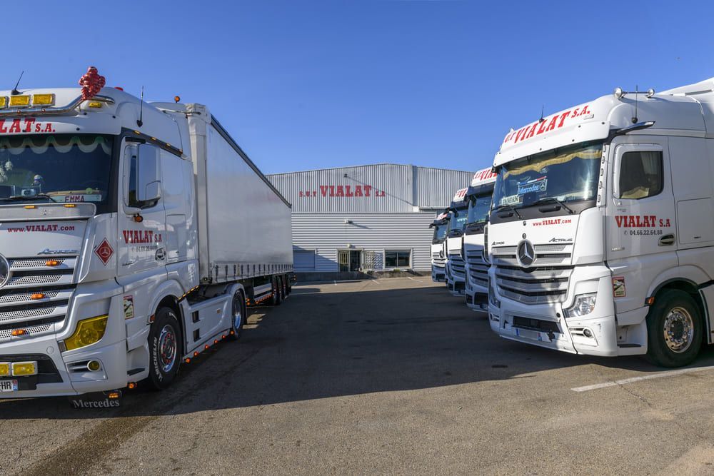 Des camions stationnés devant un entrepôt par une journée ensoleillée, avec une zone de chargement et de grandes remorques blanches.