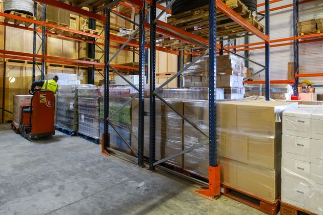 Allée d'entrepôt avec des palettes empilées, des étagères métalliques et un employé près d'un transpalette rouge