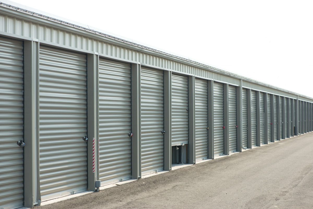 Une rangée de portes de box de stockage en métal gris longe une allée de gravier
