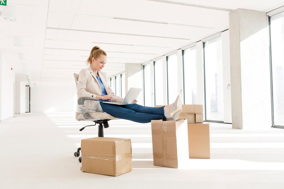 Une personne assise sur une chaise dans un couloir lumineux utilise un ordinateur portable à côté de plusieurs cartons.