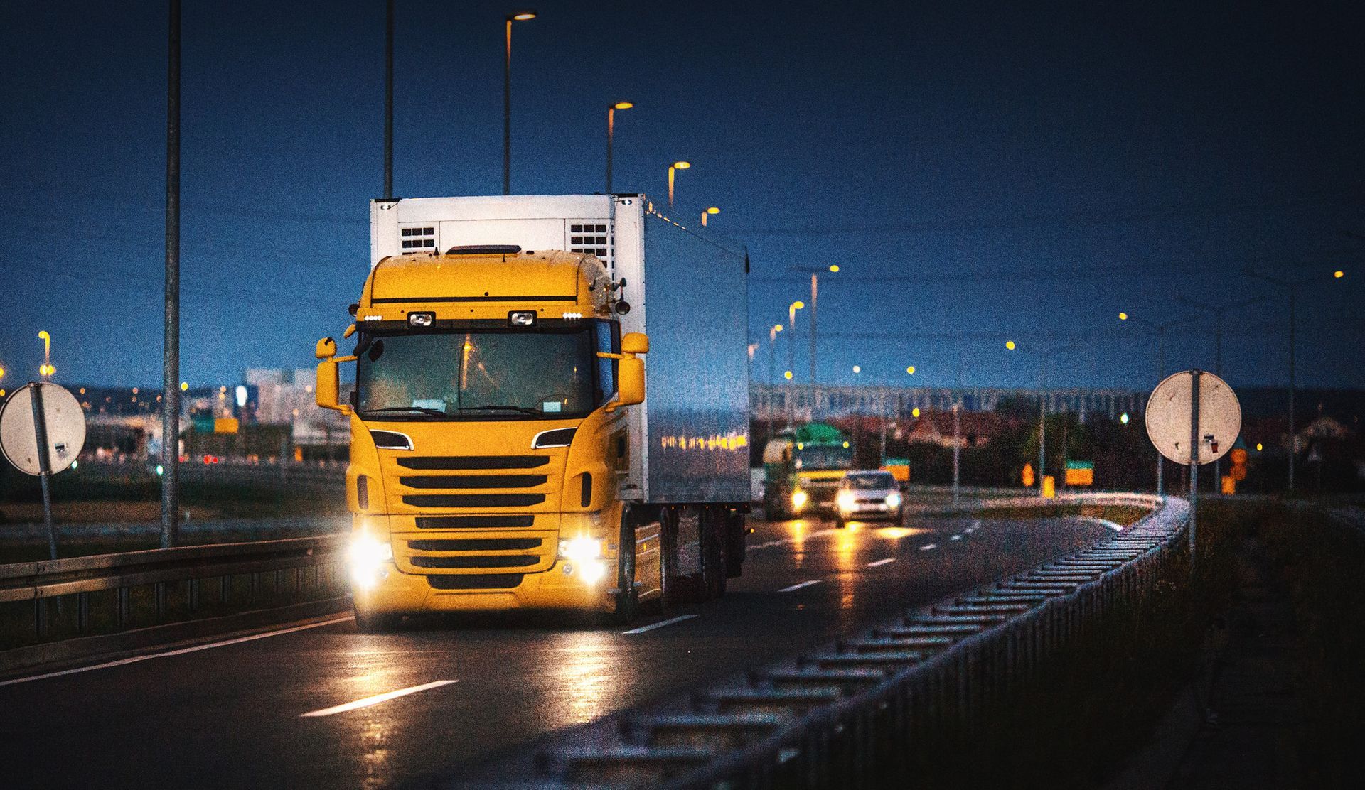 Un semi-remorque jaune circule de nuit sur une route mouillée, avec les lumières de la ville en arrière-plan.