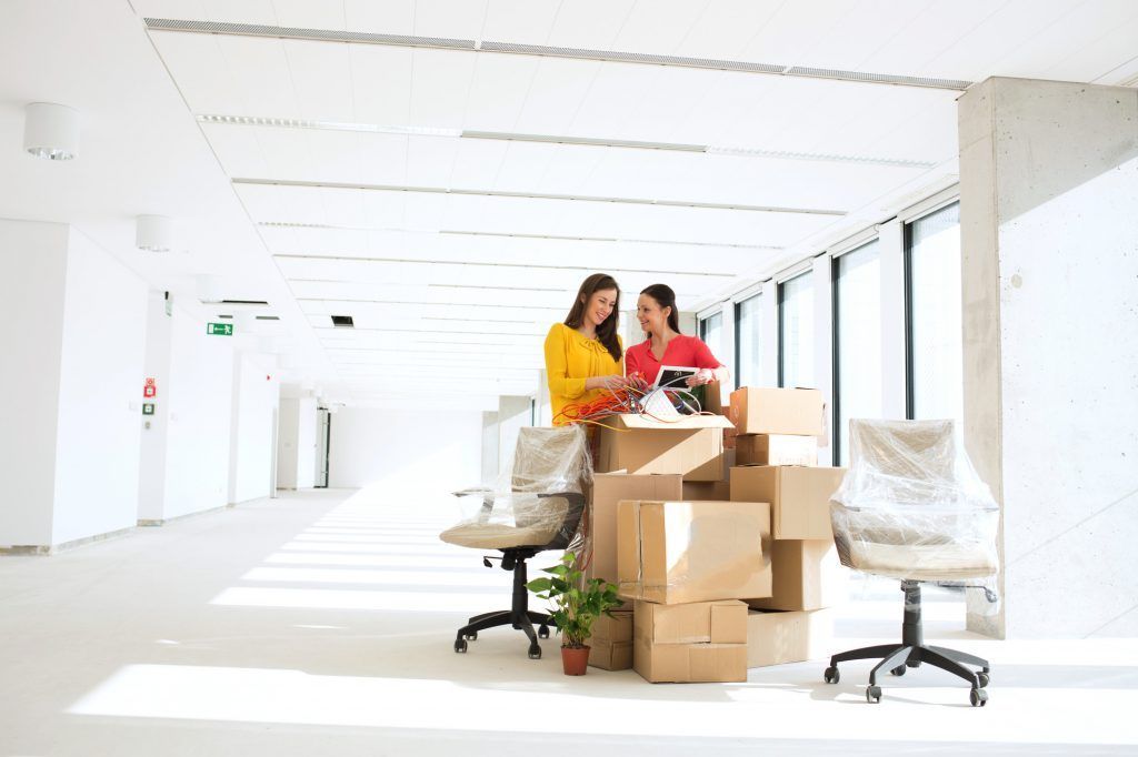 Deux personnes trient des cartons dans un bureau lumineux, près d'un bureau et de chaises.